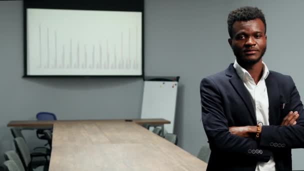 Retrato de hombre de negocios afroamericano con traje y camisa blanca de pie en la oficina en el fondo de una gran mesa y gráficos en la pantalla mirando a la cámara — Vídeo de stock