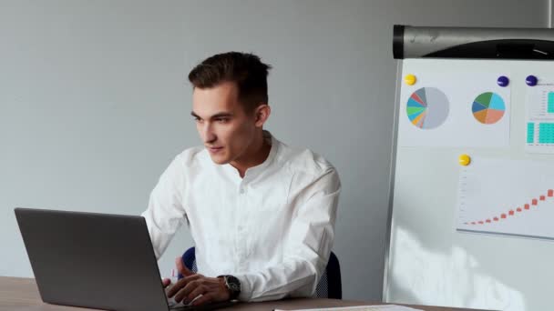 Портрет европейского мужчины, сидящего за ноутбуком в офисе в белой рубашке на фоне графиков и столов — стоковое видео