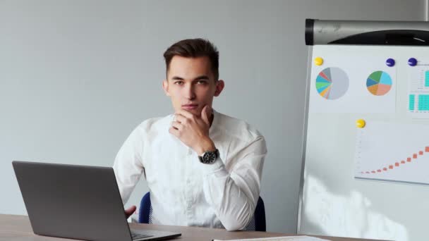 Retrato de un hombre europeo sentado en un portátil en la oficina con una camisa blanca en el fondo de gráficos y mesas. Mirando a la cámara — Vídeos de Stock