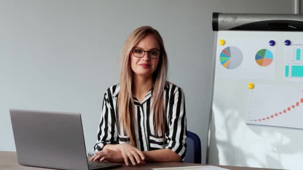 Portrait of beautiful European girl sitting at a laptop in the office with a white shirt on the background of graphs and tables. Looking at the camera — Stock Video