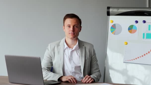 一张欧洲男性的肖像, 坐在办公室的笔记本电脑上, 上面有一件白色的衬衣, 背景是图表和桌子。看着摄像机 — 图库视频影像