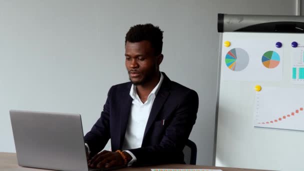 Πορτρέτο της αφρικανική αμερικανική άνθρωπος που κάθεται στο φορητό υπολογιστή στο γραφείο στο κοστούμι και λευκό πουκάμισο σε φόντο γραφήματα και πίνακες — Αρχείο Βίντεο