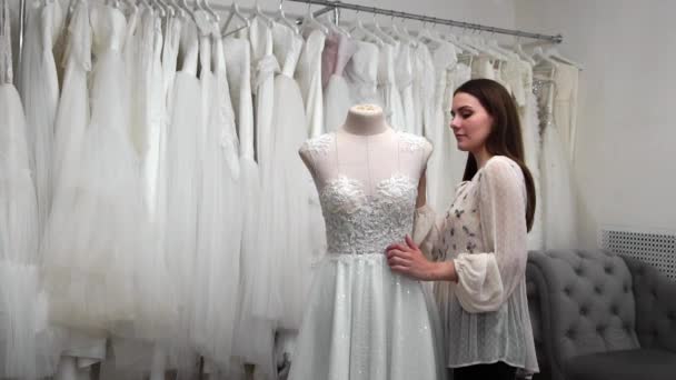 Hermosa chica buscando vestido de novia en la cabina en comparación con otros vestidos. Preparándose para la boda. Comprar un vestido de novia. La costurera inspecciona el vestido de novia terminado a medida . — Vídeos de Stock