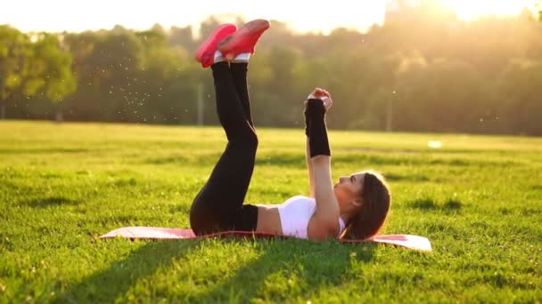 Kvinnan gör buken crunches övning på fitness mattan i sommaren park i slow motion vid solnedgången. — Stockvideo
