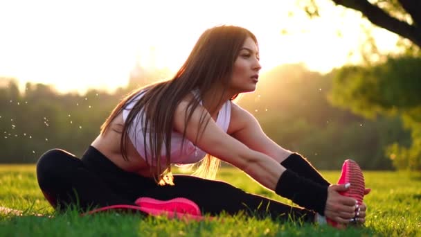 Sexy fille brune sportive en chaussures roses au ralenti au coucher du soleil dans un parc à la lumière du soleil étire les muscles des jambes avant la course du soir . — Video