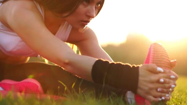 Sexy chica morena deportiva en zapatos rosados en cámara lenta al atardecer en un parque a la luz del sol estira los músculos de las piernas antes de la carrera de la noche . — Vídeo de stock