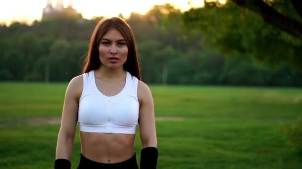 Jovem mulher bonita ouvir música no parque enquanto corre. Retrato de menina desportiva sorridente com fone de ouvido olhando para a câmera no parque no outono. Mulher atleta olhando para a câmera durante o pôr do sol de inverno . — Vídeo de Stock