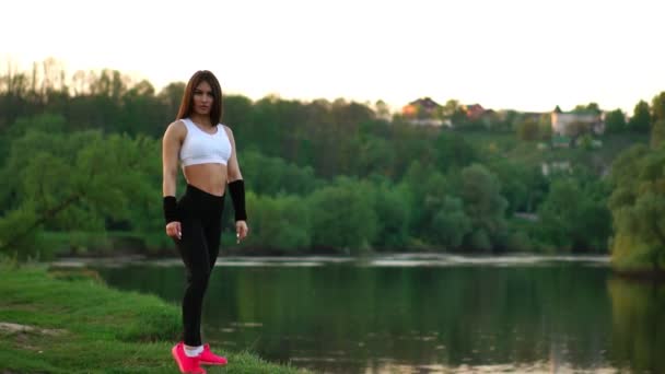 Deportiva morena en un top blanco pantalones ajustados negros y zapatillas de deporte de color rosa en la luz del sol estira los músculos y se prepara para el entrenamiento en el aire fresco en el campo sobre el fondo del bosque . — Vídeos de Stock