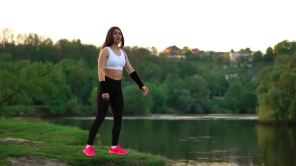 Sportovní bruneta v bílém horní černé těsné kalhoty a růžové tenisky v slunci úseky svaly a připraví na výcvik na čerstvém vzduchu v poli pozadí lesa. — Stock video