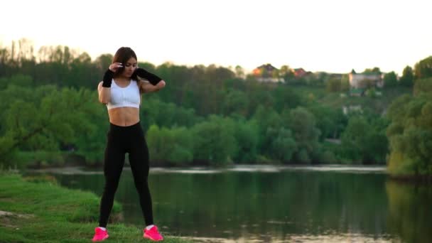 Deportiva morena en un top blanco pantalones ajustados negros y zapatillas de deporte de color rosa en la luz del sol estira los músculos y se prepara para el entrenamiento en el aire fresco en el campo sobre el fondo del bosque . — Vídeos de Stock