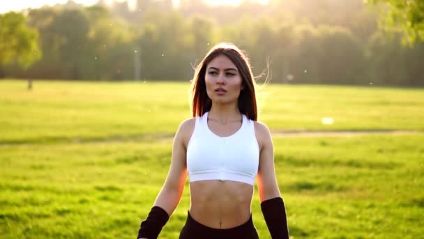 Brune sportive dans un pantalon serré noir haut blanc et baskets roses dans la lumière du soleil étire les muscles et se prépare à l'entraînement dans l'air frais dans le domaine sur le fond de la forêt . — Video