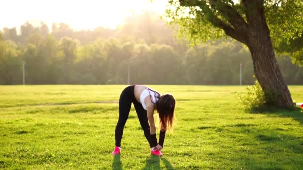 Спортивная брюнетка в белых черных узких брюках и розовых кроссовках на солнце растягивает мышцы и готовится к тренировкам на свежем воздухе в поле на фоне леса . — стоковое видео