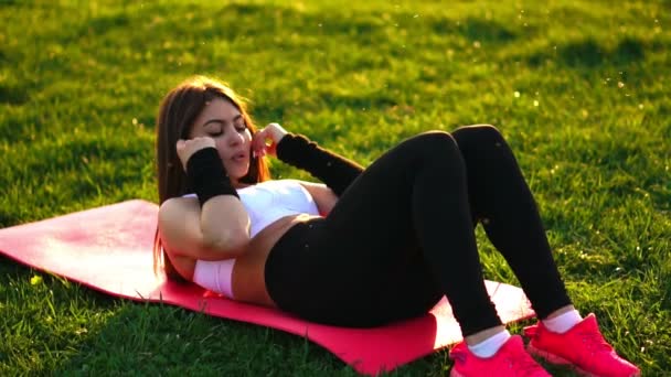 Женщина делает упражнения на живот на коврике для фитнеса в летнем парке в замедленной съемке на закате . — стоковое видео