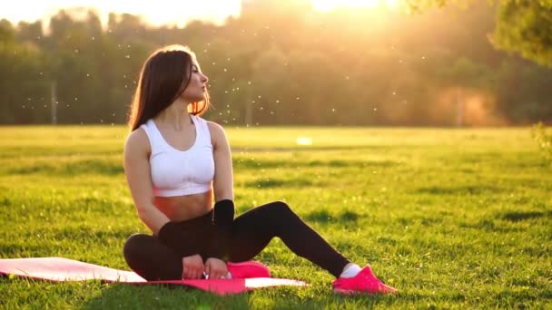 Mujer joven y hermosa está sentada en la alfombra durante el descanso en su entrenamiento de fitness. Descanse después del ejercicio a la luz del sol corrigiendo el cabello en cámara lenta. Cabello en la luz de fondo . — Vídeo de stock