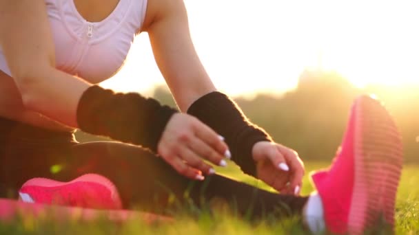 Le mani ravvicinate prendono il sopravvento sulla suola delle sneakers sull'erba alla luce del sole. Scaldarsi prima della corsa mattutina. Bella bruna su una corsa nel parco . — Video Stock