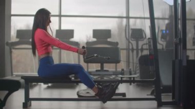 Spor salonunda yoğun güç eğitimi. Oturan bir kadın, bir simülatörün ağırlığını büyük pencerelerin ve spor salonunun koşu bandının arka planına doğru çekiyor.