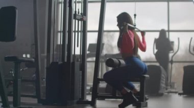 Genç bir kadın onu spor salonunda çalıştırıyor. Arka planda geniş pencereleri olan bir odada eğitim gören bir kadın elipsoid ve koşu bandı kullanır.