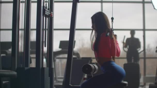 Gadis itu menggoyangkan punggungnya saat latihan. Pelatihan di sebuah ruangan dengan Windows besar di latar belakang seorang wanita menggunakan elipsoid dan treadmill — Stok Video