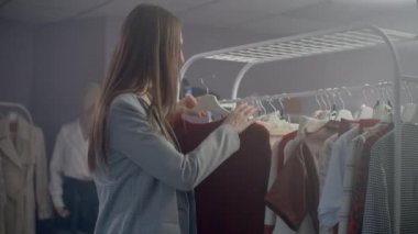 Giyim mağazasındaki genç bir esmer askıya asmak için bir elbise seçer ve onu dener. Mağazadan bir elbise al. Bir butikte elbise ara ve seç..