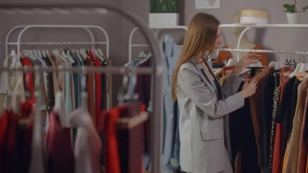 Een jonge Brunette in een kledingwinkel kiest een jurk om op te hangen en probeert het. Koop een jurk in de winkel. Zoek en kies kleding in een boetiek. — Stockvideo