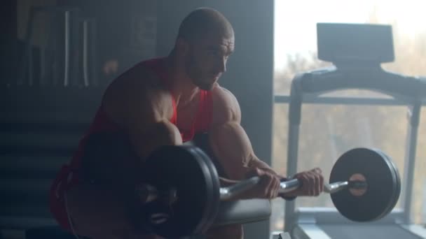 En stark manlig kroppsbyggare lyfter en skivstång när han sitter på bänken och tränar hårt i slow motion på gymmet — Stockvideo