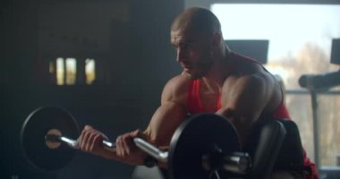 Erkek sporcu ağırlık kaldırarak bir bankta oturuyor. Yavaş hareket eden eller.
