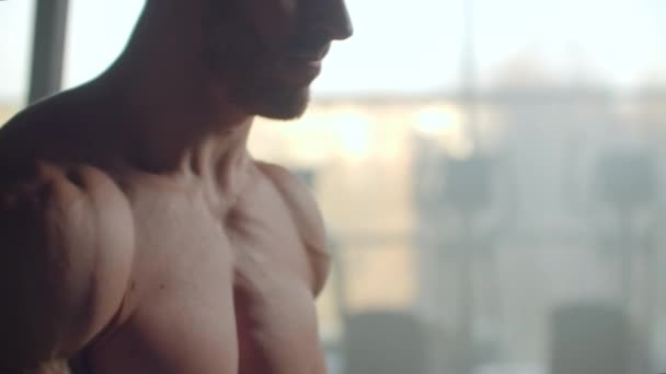 Un hombre con un torso desnudo y un cuerpo fuerte bombeado y los músculos de alivio de la prensa levanta una barra con un peso en el fondo de la ventana entrenando sus manos . — Vídeos de Stock