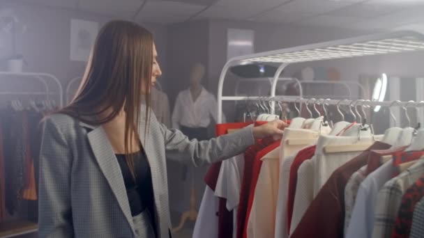 Una giovane bruna in un negozio di abbigliamento sceglie un abito da appendere a un gancio e lo prova. Compra un vestito nel negozio. Cerca e scegli i vestiti in una boutique . — Video Stock