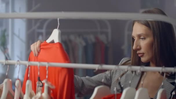 En ung Brunett i en klädbutik väljer en klänning att hänga på en Hanger och provar den på. Köp en klänning i butiken. Sök och välj kläder i en butik. — Stockvideo
