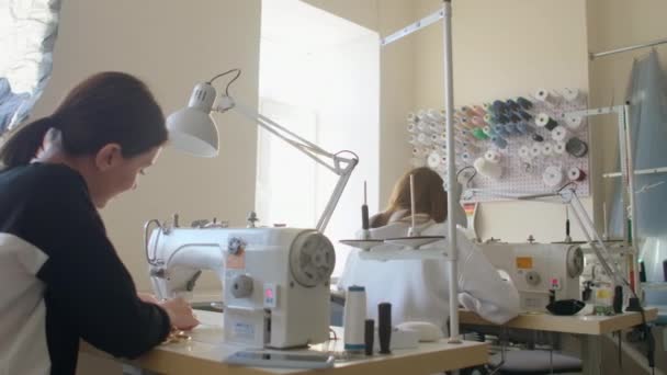 Két varrónő varrógépeken dolgozik a ruhadarab szabóműhelyében. Tervezői ruhák készítése. Kisvállalkozás. Két alkalmazott dolgozik varrógépeken hátulról.. — Stock videók