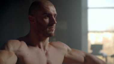 Tişörtsüz bir adam pencerenin önündeki dambılları kaldırıyor omuz kasları için egzersiz yapıyor, vücut geliştirme eğitimi alıyor. Sağlıklı yaşam tarzı ve kas kütlesi kazanmak için egzersizler. Kuvvet eğitimi 