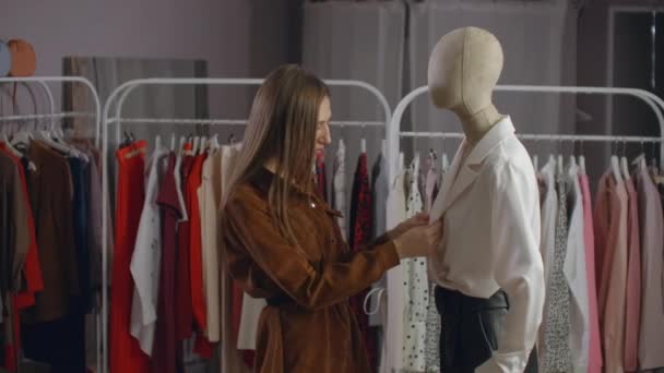 Une belle fille dans une veste regarde les vêtements sur un mannequin, touche et considère le style et le type de tissu de vêtements dans la salle d'exposition — Video
