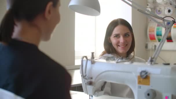 A fiatal nő varrógéppel dolgozik, és ellenőrzi a varratokat, amikor a kollégája jön hozzá vázlattal. Nők. Szövet és beszéd.. — Stock videók