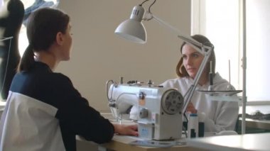 Genç kadın dikiş makinesiyle çalışıyor ve iş arkadaşı çizim için geldiğinde dikişleri kontrol ediyor. Kadınlar işte. Kumaşa bakıyor ve konuşuyor..