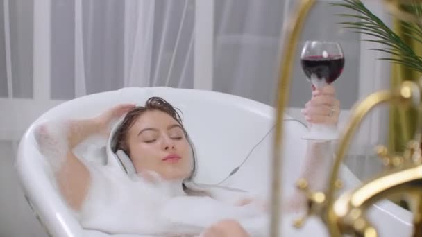 Mulher morena sexy Ouvindo música no banho e bebendo vinho, relaxando e relaxando no banho de espuma deitado. Tome um banho depois de uma semana difícil. Recuperar — Vídeo de Stock