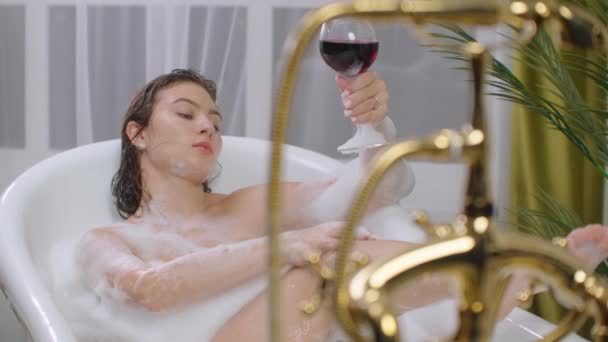 Beautiful young brunette woman enjoying pleasant bath with foam, lying with closed eyes and holding a glass of wine. — Stock Video