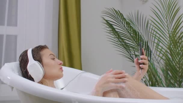 한 아름다운 갈색의 여성 이 스마트폰으로 음악을 듣고 있습니다. 그 여성은 거품이 섞인 욕조에 누워 근무일로부터 쉬고 있습니다. — 비디오