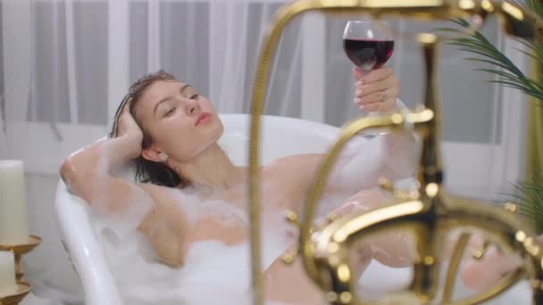 Жінка п'є вино в ванні з піною. Молода мила дівчина п'є червоне вино у ванні з піною. Вона відпочиває після важкого дня. Дівчина спокійно дивиться вгору і відправляє — стокове відео