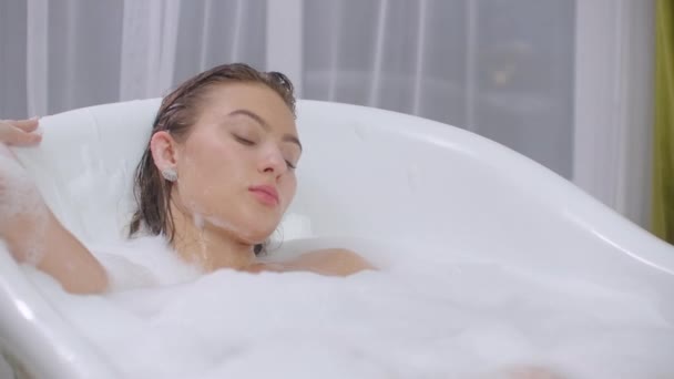 Schöne Frau entspannt sich im Schaumbad liegend in der Badewanne. fröhlich und entspannt in der Badewanne liegend mit Schaumbad zu Hause oder im Hotelbadezimmer lächelnd spielerisch Luxus genießen — Stockvideo