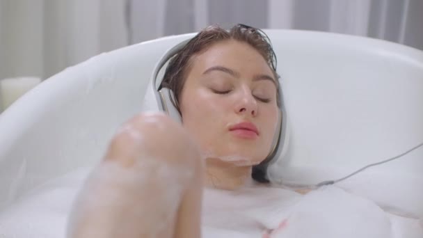 Écouter de la musique dans un bain moussant. Jeune brune se détend dans la salle de bain — Video