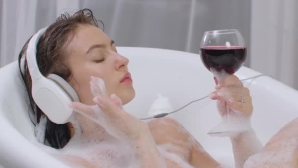 Sıcak bir köpük banyosunda uzanmış kırmızı şarap iç ve rahatlamak için müzik dinle. — Stok video