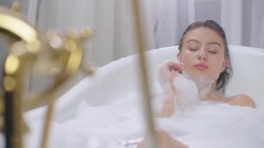 Küvette uzanmış köpük banyosunda dinlenen güzel bir kadın. Neşeli ve rahat bir şekilde küvette uzanmış köpük köpükler içinde evde ya da otel banyosunda gülümsüyor ve lüks yaşam tarzının keyfini çıkarıyordu.