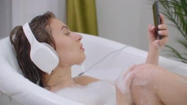 Küvette müzik dinleyen güzel bir kadın rahatlatıcı köpük banyosu yaşam tarzının tadını çıkarıyor..