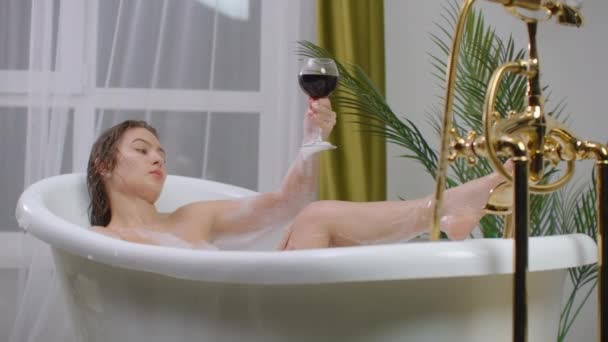 아름답고 젊은 갈색의 여자가 거품 목욕을 하면서 편안하게 휴식을 취하고 붉은 포도주를 마신다. 따뜻 한 거품 목욕으로 건강을 회복하고 긴장을 풀라 — 비디오