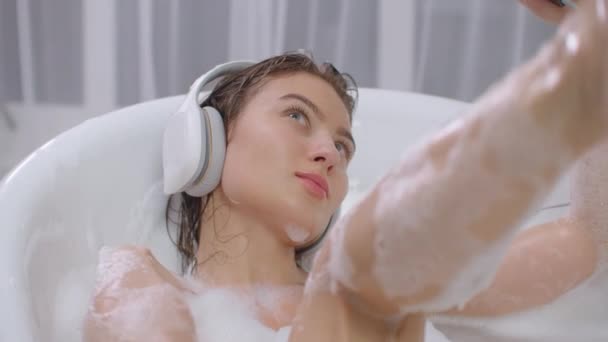 Połóż się w łazience z pianką i zrelaksuj słuchając muzyki przez białe słuchawki. Relaks i relaks w wannie — Wideo stockowe