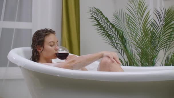 En förförisk brunett ligger i ett bad med skum som slappnar av och dricker rött vin. Återhämta dig och slappna av i ett varmt bubbelbad — Stockvideo