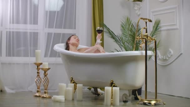 Egy gyönyörű fiatal barna nő fekszik egy pezsgőfürdőben pihentető és vörös bort iszik. Gyógyuljon meg és lazítson egy forró habfürdőben. — Stock videók