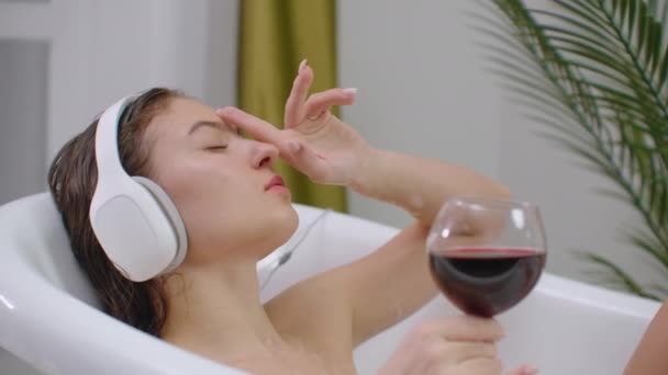 Una joven hermosa morena caucásica yace en el baño y escucha música en auriculares blancos descansando del estrés y relajándose bebiendo vino tinto de una copa . — Vídeo de stock