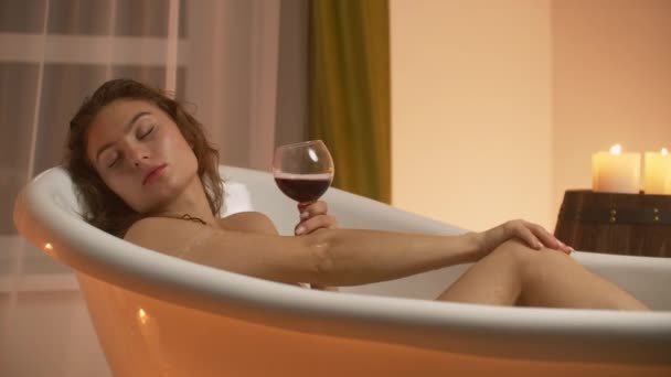 Eine junge schöne kaukasische Brünette liegt im Badezimmer bei Kerzenlicht in einer angenehmen Abendatmosphäre, die sich vom Stress ausruht und entspannt Rotwein aus einem Glas trinkt. — Stockvideo