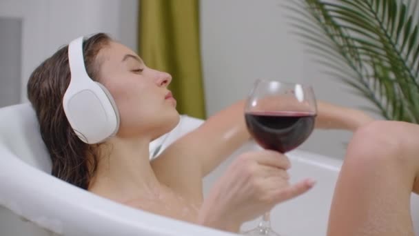 Relaxou jovem ouvindo música em uma bolha ba. Close-up de uma jovem relaxada ouvindo música com fones de ouvido em um banho de espuma e beber vinho tinto de um copo . — Vídeo de Stock